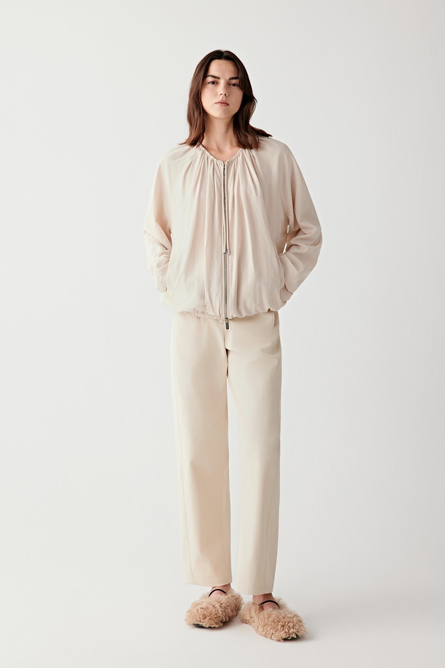 Full-length front view of a woman wearing light beige stretch cotton gabardine trousers with a straight cut and high waist, showing clean lines and structured drape