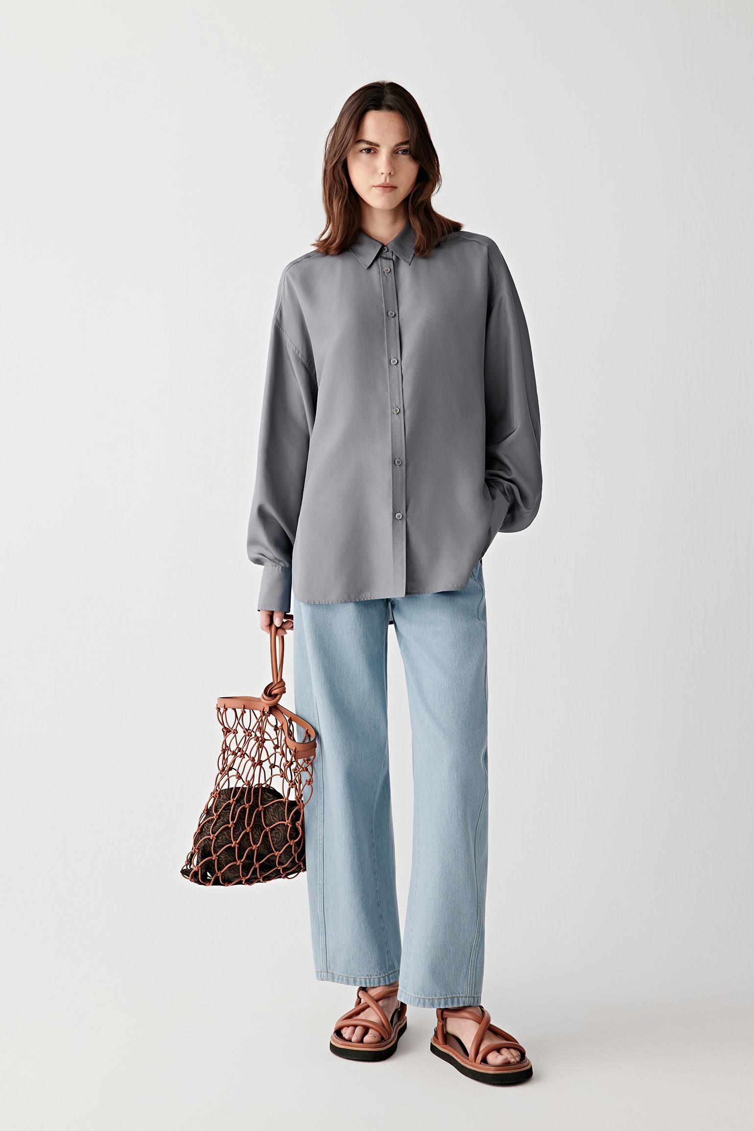 Woman wearing a gray shirt and light blue pants, holding a woven bag on a white background