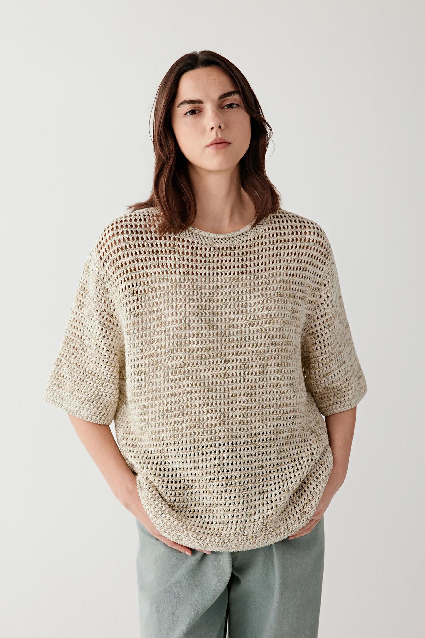 Three-quarter front view of a woman wearing a beige open-knit cotton pullover with a relaxed fit, short sleeves and airy structure, paired with loose trousers