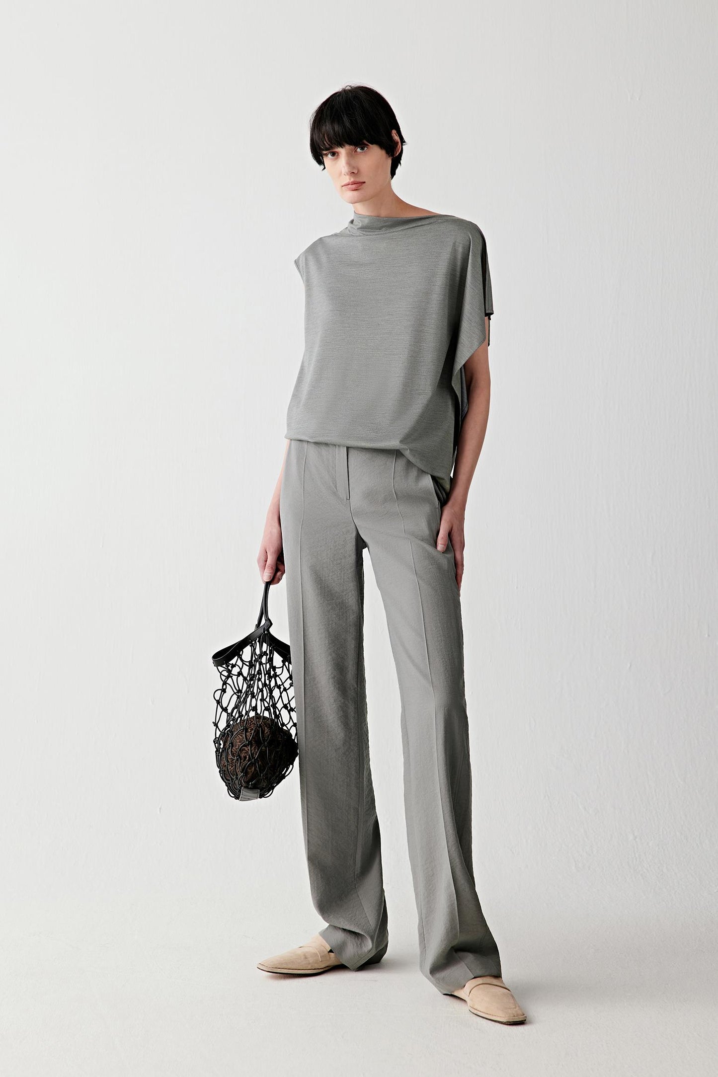 Full-length front view showing the relaxed silhouette shaped by a draped collar, open shoulders and asymmetrical sleeves. The stretch jersey construction follows the movement of the body while maintaining a soft and fluid line.