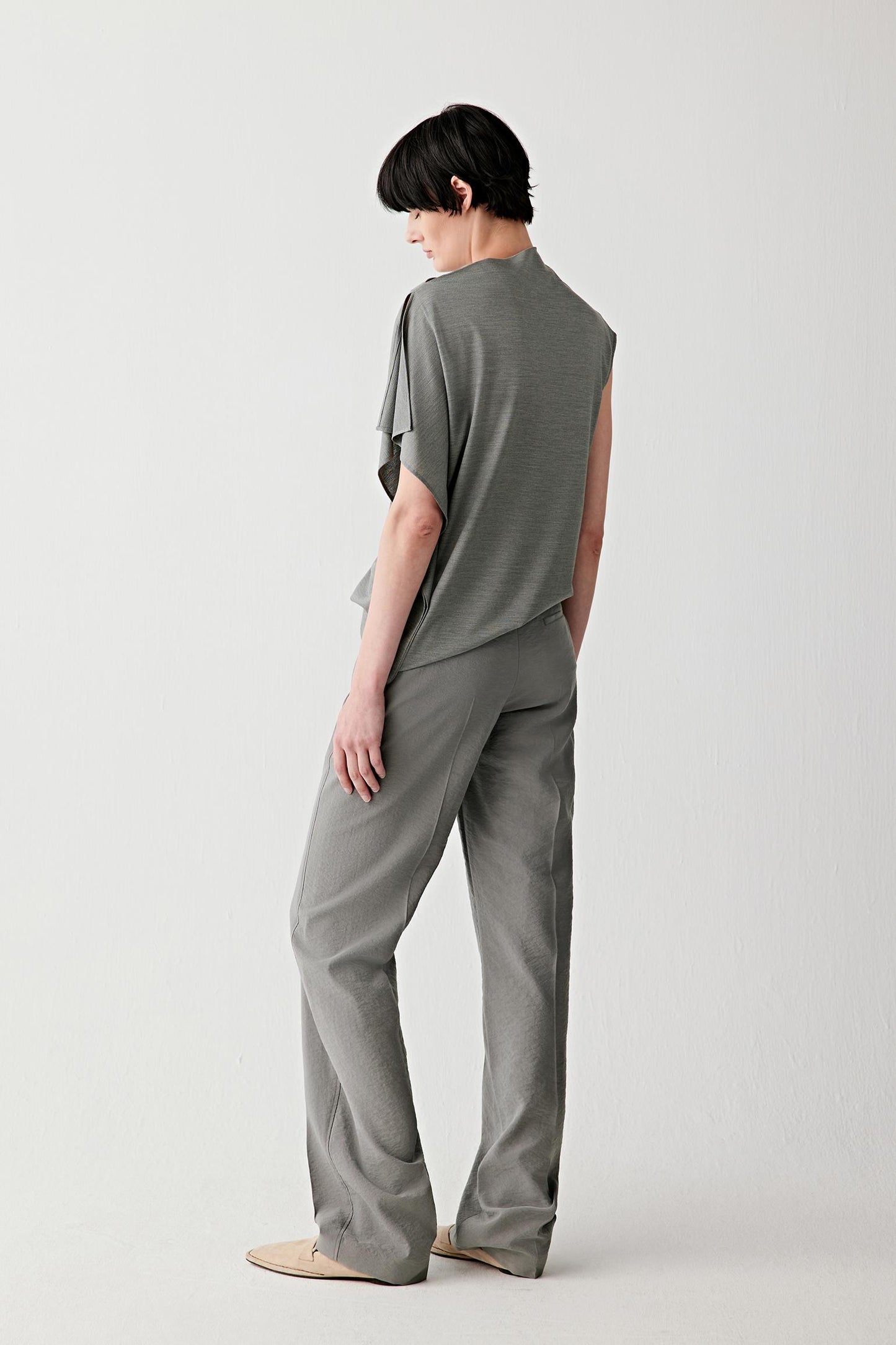 Side or three-quarter back view emphasizing the elongated drape and sculptural quality of the silhouette. The fluid jersey falls softly through the body, while the draped neckline creates movement and depth around the upper construction.