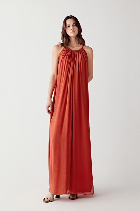 Front view of a woman wearing a deep red long dress in silk mousseline, highlighting the lightweight texture, fluid drape and softly translucent movement of the fabric. The surface reveals subtle tonal variations across the flowing silhouette.