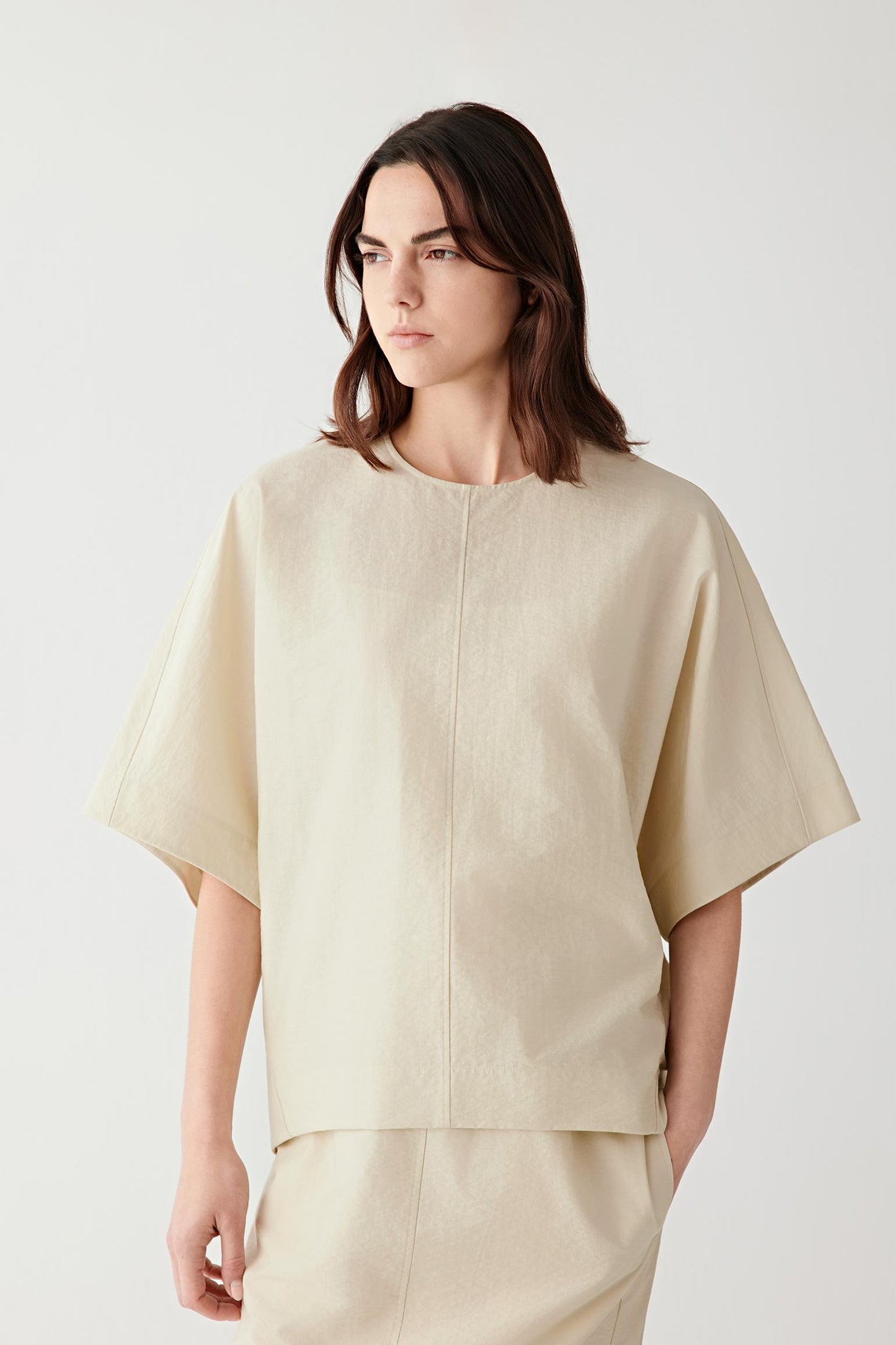 Front view of a woman wearing a light beige top in cotton and nylon crinkled poplin, highlighting the lightweight crinkled texture, fluid drape and softly irregular surface