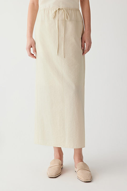 Front view of a woman wearing a light cream skirt in crinkled cotton and nylon poplin, highlighting the lightweight construction, softly textured surface and fluid straight silhouette. The fabric combines a dry hand with subtle irregularity, creating gentle tonal variation across the garment.