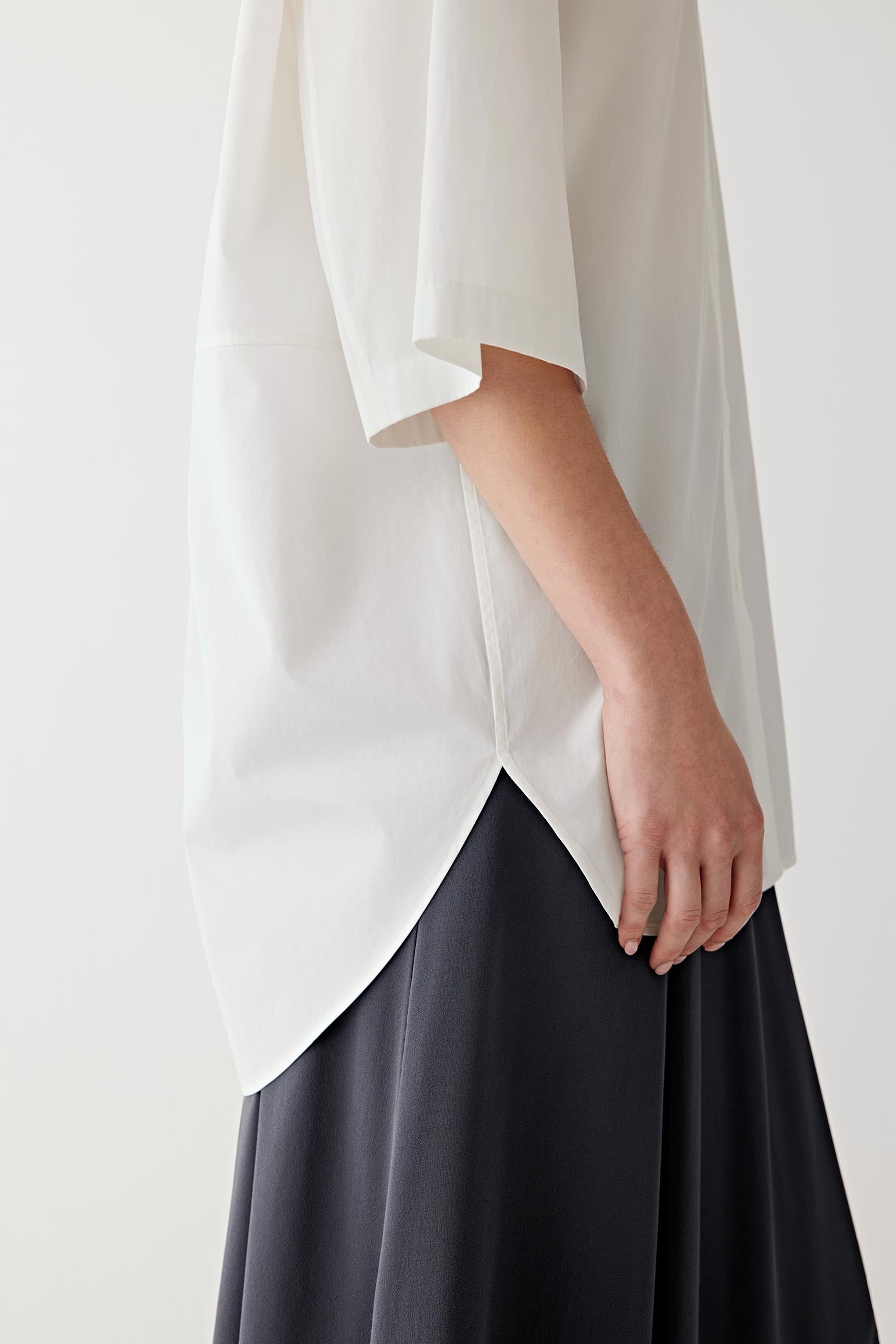 Close-up side view of the curved hem and side slit of a white cotton poplin shirt, highlighting the crisp smooth surface, clean finishing and lightweight structured drape of the fabric against a fluid dark skirt silhouette