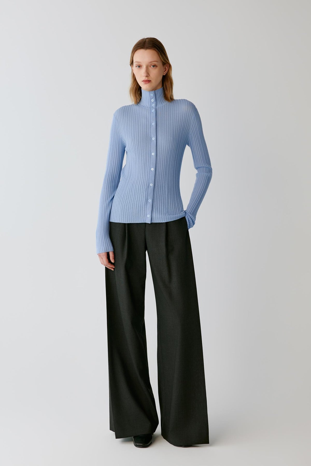 A model stands against a light gray studio background wearing a light blue ribbed long-sleeve button-front top with a high mock neck and black wide-leg trousers.