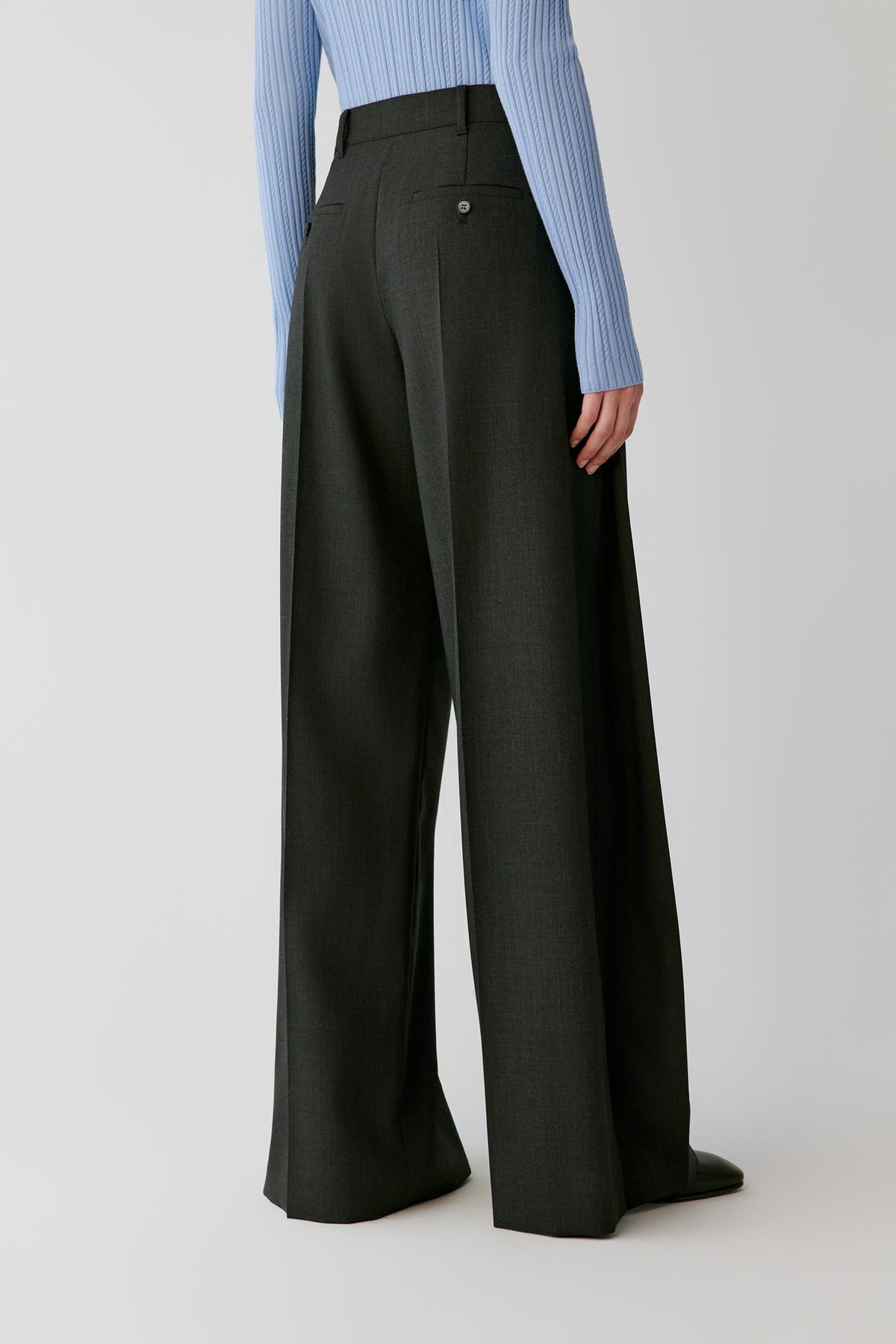 A model from the back wearing high-waisted, wide-leg charcoal gray trousers and a light blue ribbed long-sleeve top, photographed against a neutral light gray studio background.