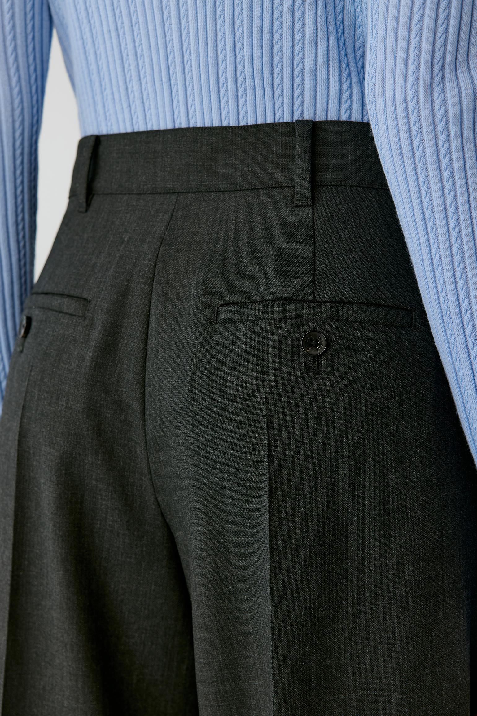 Close-up of dark charcoal gray trousers shown on a model, featuring a buttoned welt pocket and belt loops, paired with a light blue ribbed knit sweater.
