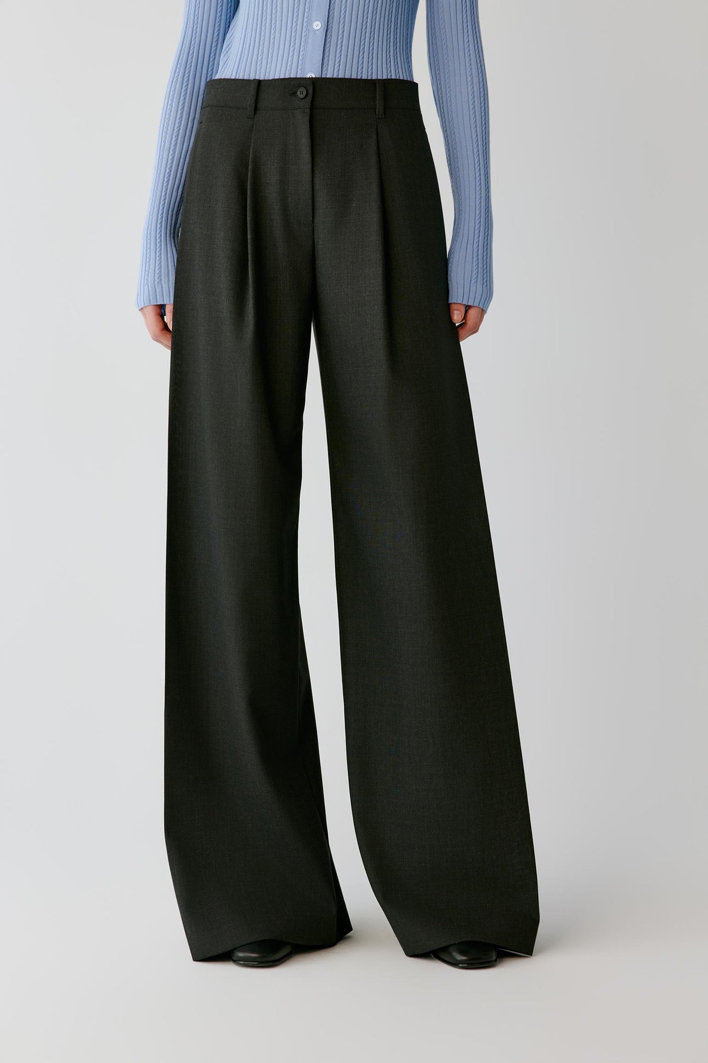 A model wearing black wide-leg trousers with a button closure and front pleats, styled with a light blue ribbed long-sleeve shirt and black shoes, against a light gray studio background.