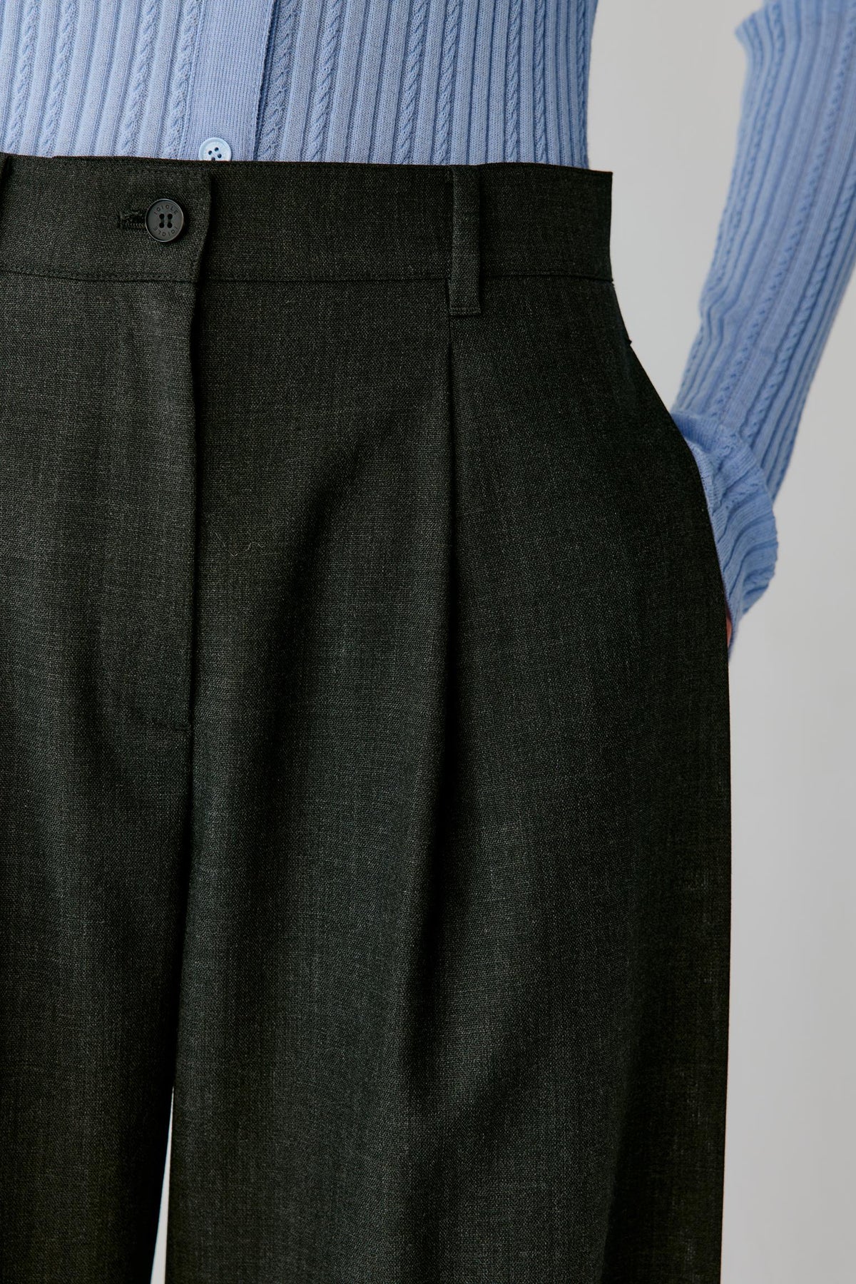 Close-up of dark charcoal gray pleated trousers worn with a light blue ribbed knit top, showing the waistband, button, and fabric texture.