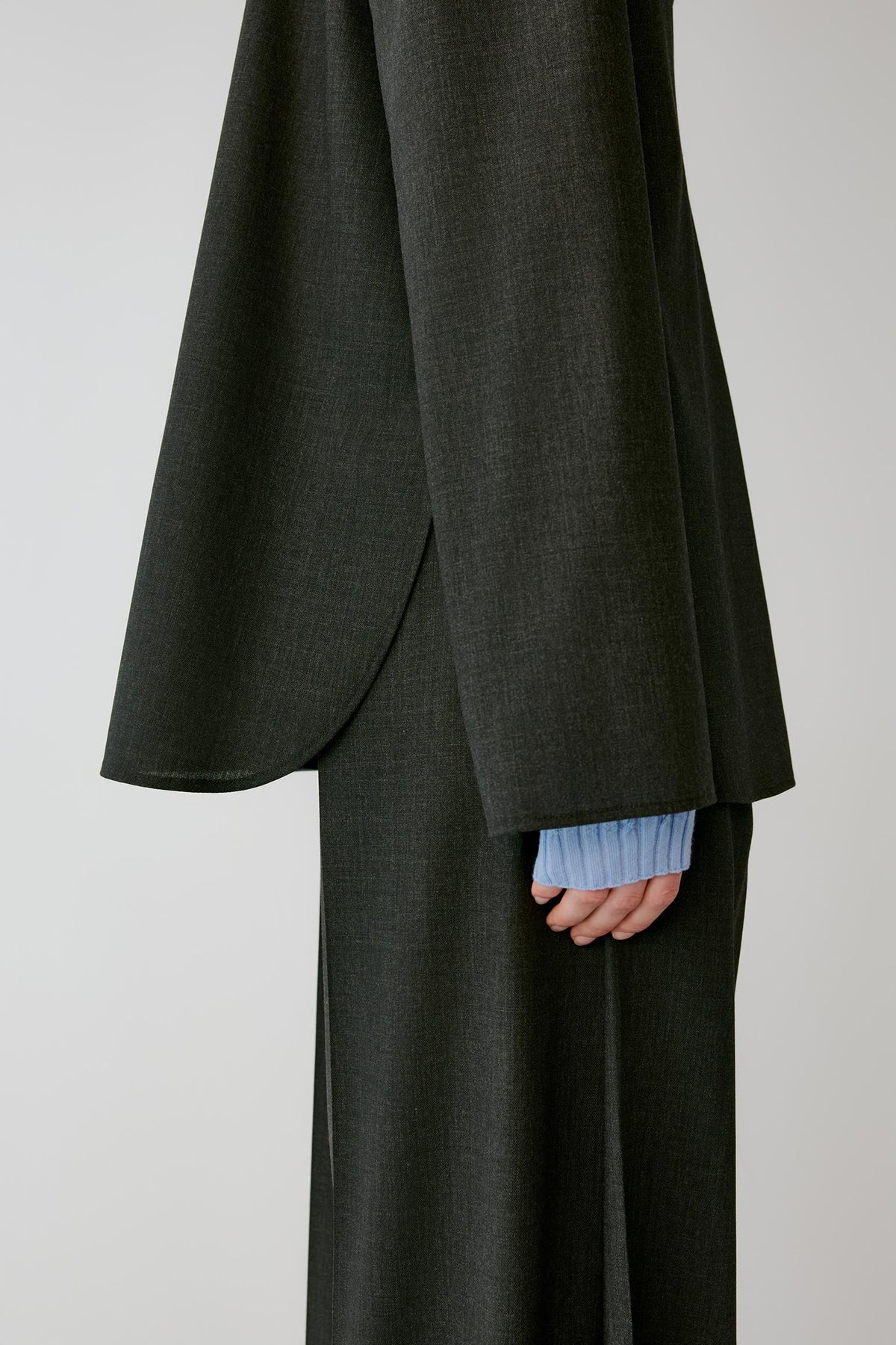 A close-up of a person wearing a dark charcoal gray draped jacket or kimono-style top with wide flowing sleeves, paired with matching wide-leg trousers. A light blue ribbed shirt cuff is visible at the hem.