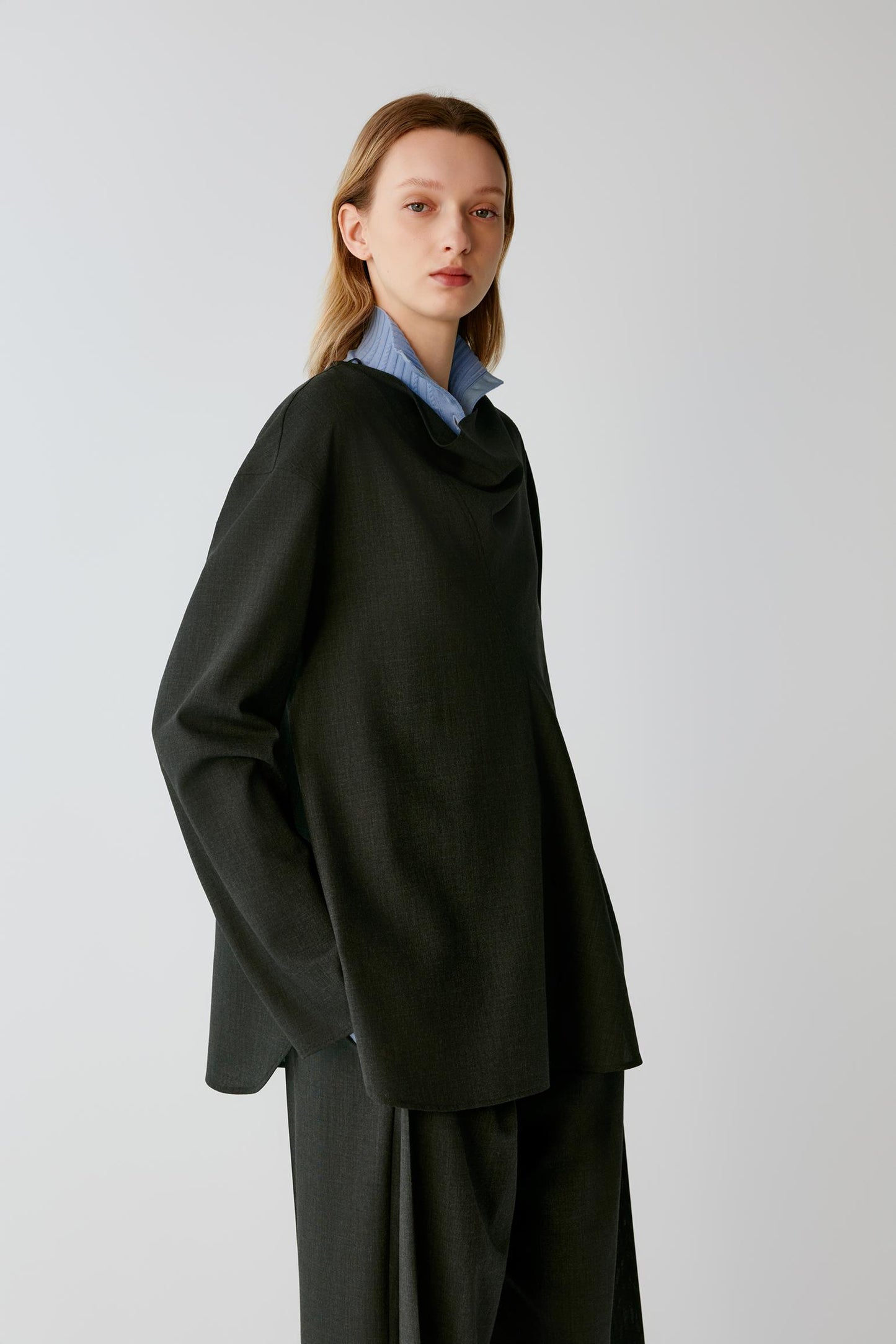 A model wearing an oversized black draped cowl-neck top and matching wide-leg trousers, layered over a light blue striped collared shirt, against a plain light gray studio background.