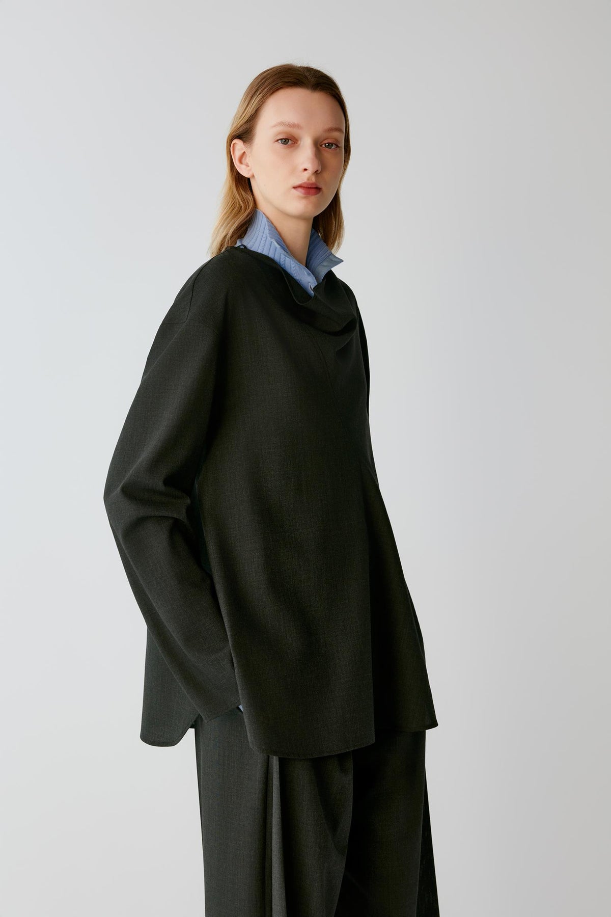 A model wearing an oversized black draped cowl-neck top and matching wide-leg trousers, layered over a light blue striped collared shirt, against a plain light gray studio background.
