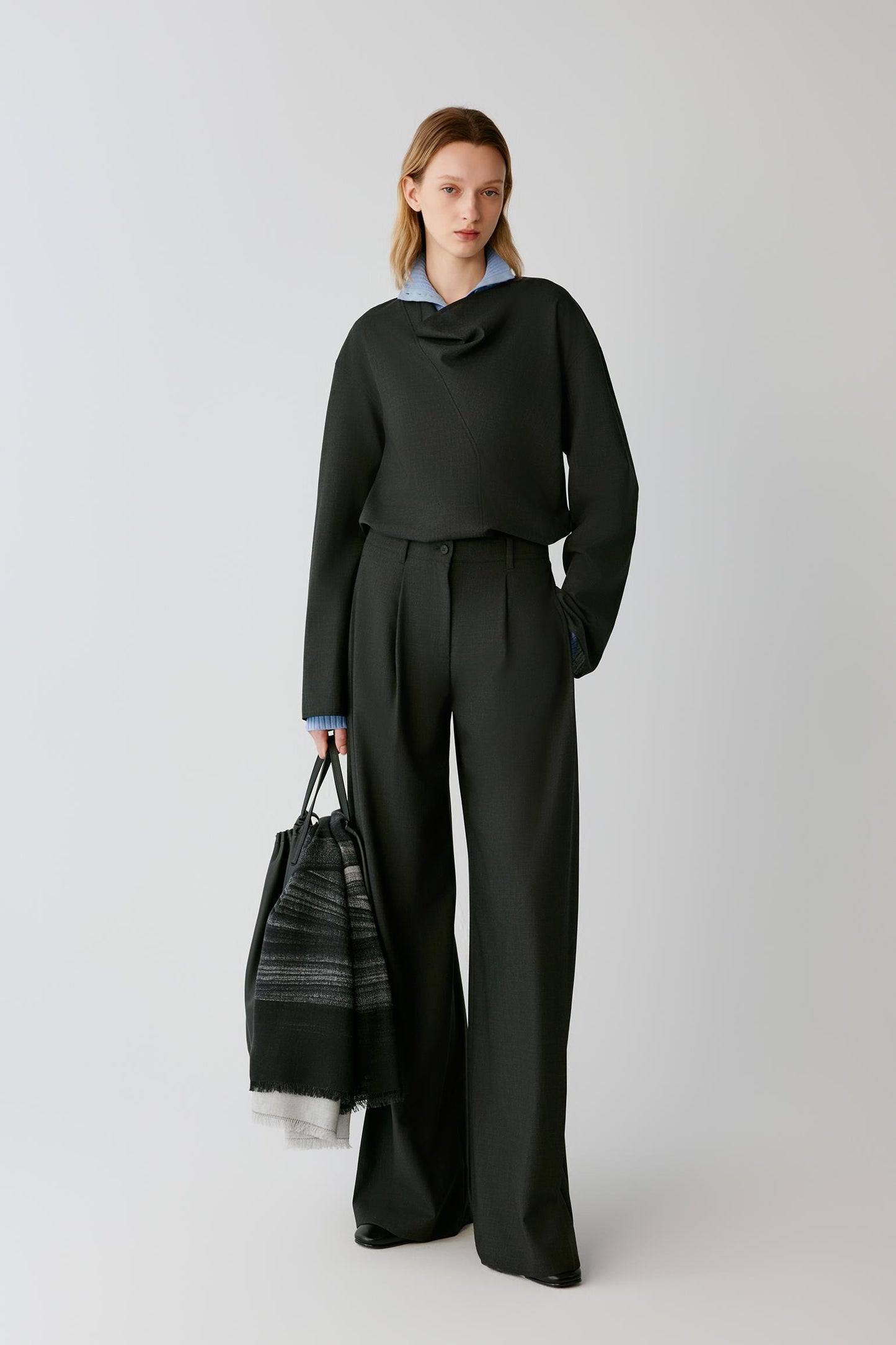 A model stands against a light gray studio background wearing a dark charcoal gray draped long-sleeve top and matching wide-leg trousers, with a light blue collared shirt peeking out at the neckline. She holds a black and gray patterned tote bag.