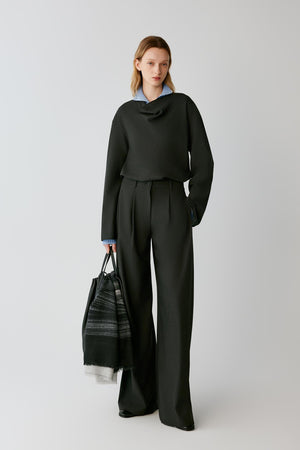 A model stands against a light gray studio background wearing a dark charcoal gray draped long-sleeve top and matching wide-leg trousers, with a light blue collared shirt peeking out at the neckline. She holds a black and gray patterned tote bag.