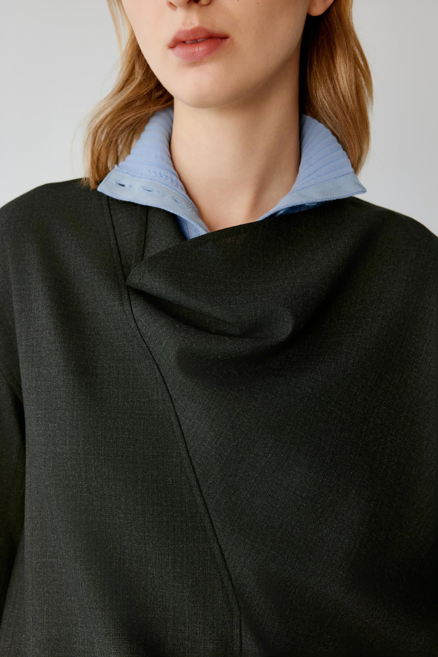 Close-up of a model wearing a dark charcoal gray wrap-style top with a draped cowl neckline, layered over a light blue collared shirt.