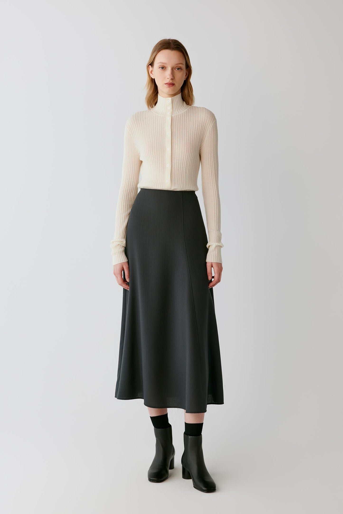 A model stands against a light gray studio background wearing a cream ribbed mock-neck long-sleeve top, a black midi skirt, black socks, and black ankle boots.