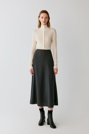 A model stands against a light gray studio background wearing a cream ribbed mock-neck long-sleeve top, a black midi skirt, black socks, and black ankle boots.