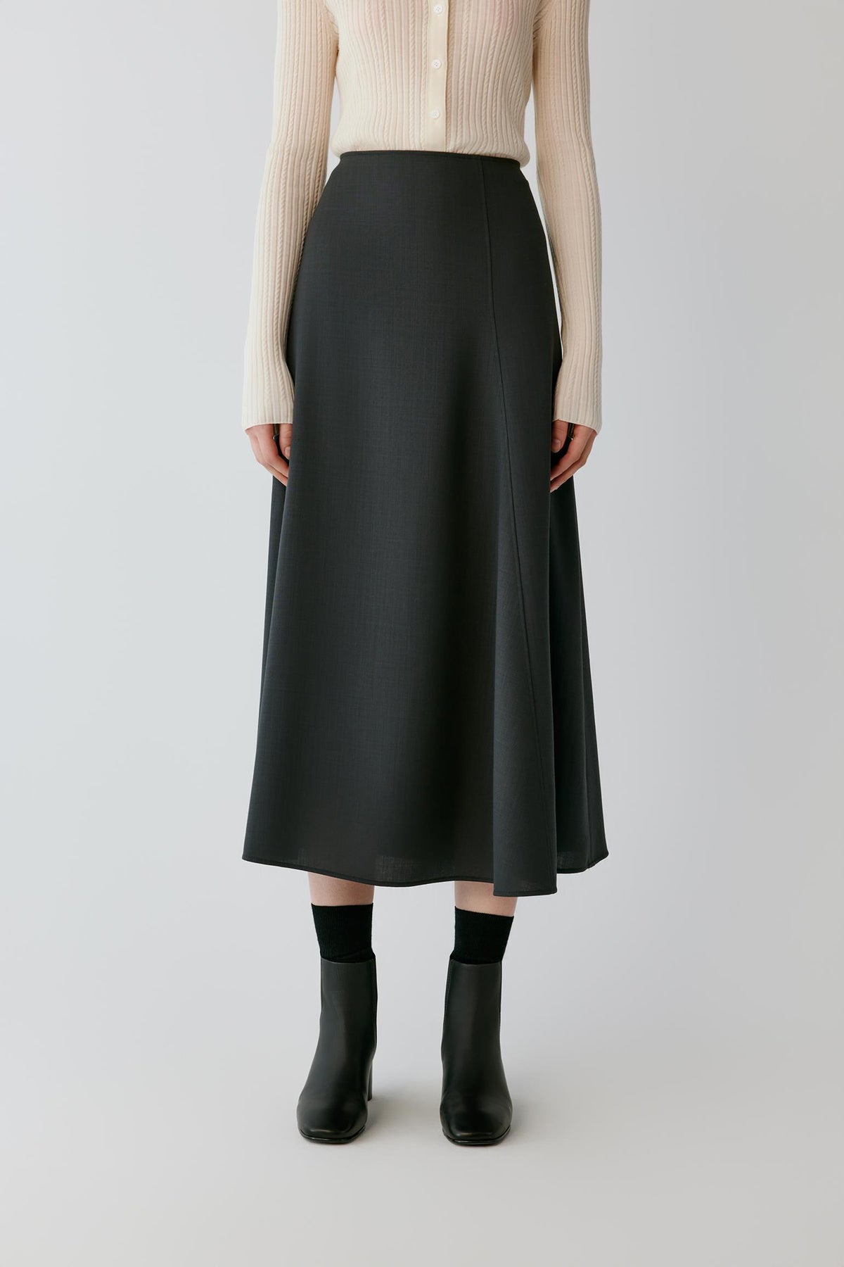 A model wearing a black midi skirt with a subtle vertical texture, paired with a cream ribbed cardigan, black socks, and black ankle boots, shot against a light gray studio background.
