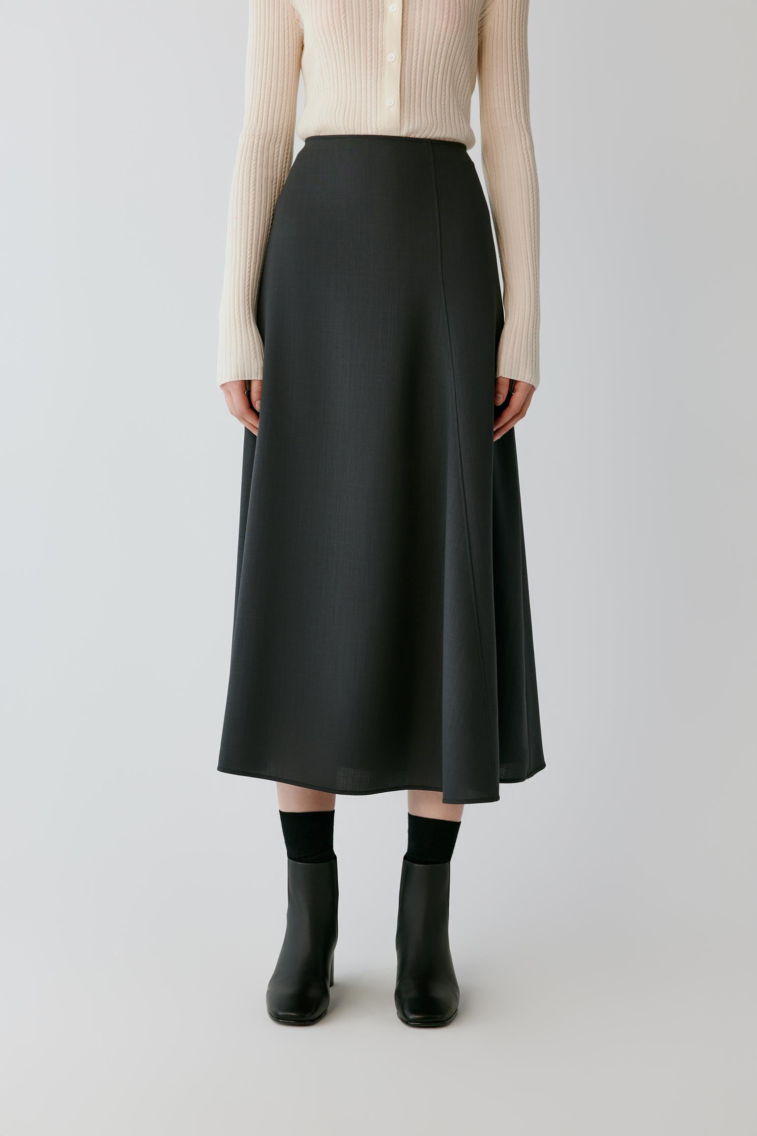 A model wearing a black midi skirt with a subtle vertical texture, paired with a cream ribbed cardigan, black socks, and black ankle boots, shot against a light gray studio background.