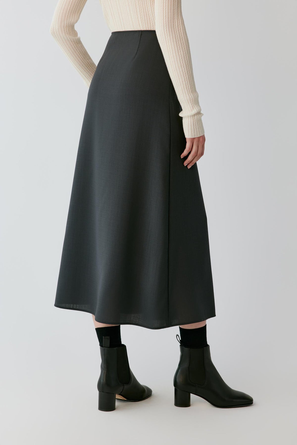 A model is shown from the back wearing a dark charcoal gray A-line midi skirt, a cream ribbed long-sleeve top, and black ankle boots with gold-tone heels, set against a light gray studio background.