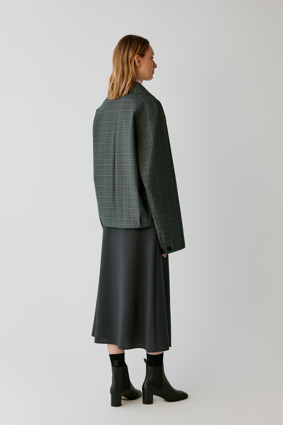 A model is shown from the back wearing a dark green plaid blazer, a black midi skirt, and black ankle boots against a light gray studio background.