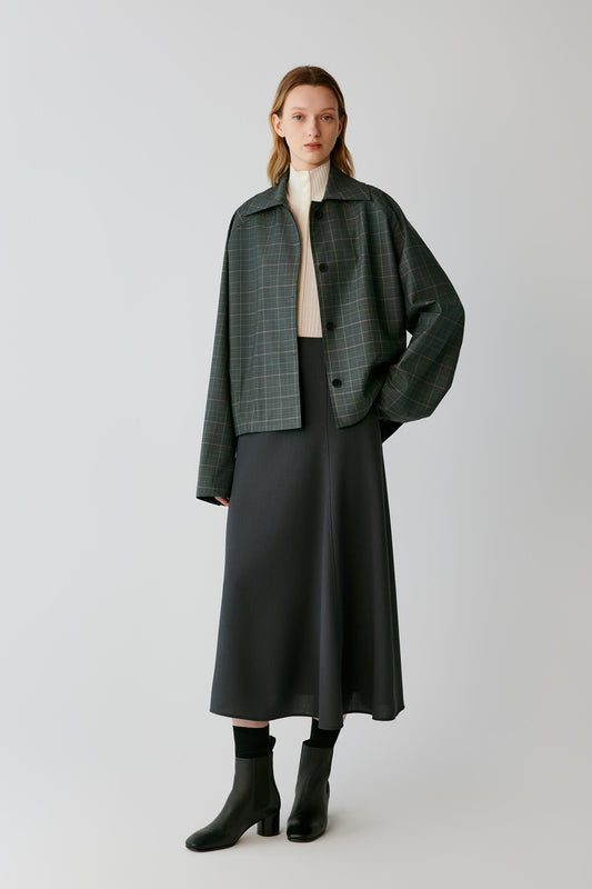 A model stands against a light gray studio background wearing a dark green and black plaid cropped jacket over a cream turtleneck, paired with a long black skirt and black ankle boots.