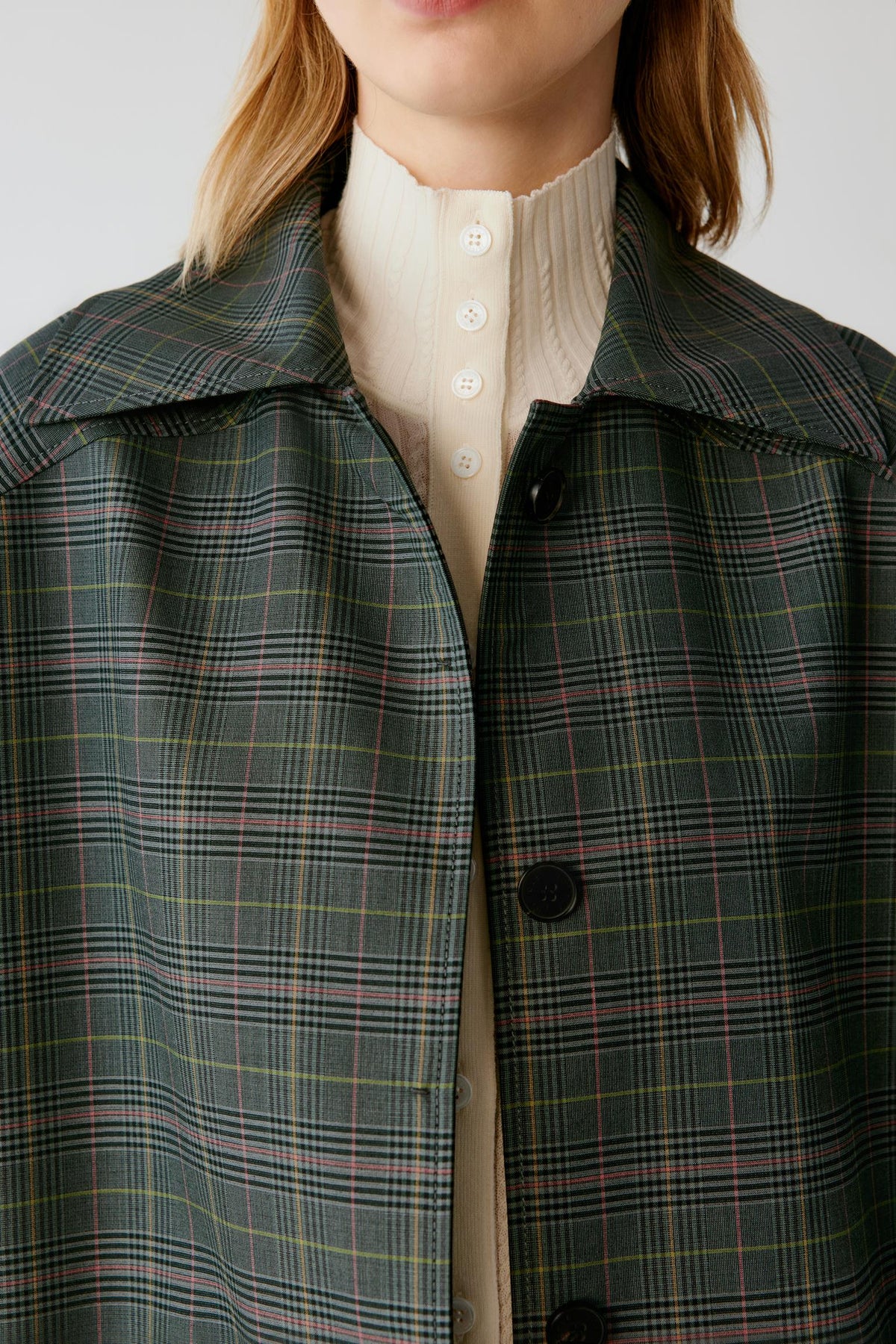 A close-up of a model wearing a dark green plaid coat over a cream ribbed turtleneck sweater. The coat features a wide collar, black buttons, and a subtle plaid pattern with thin yellow and pink lines.