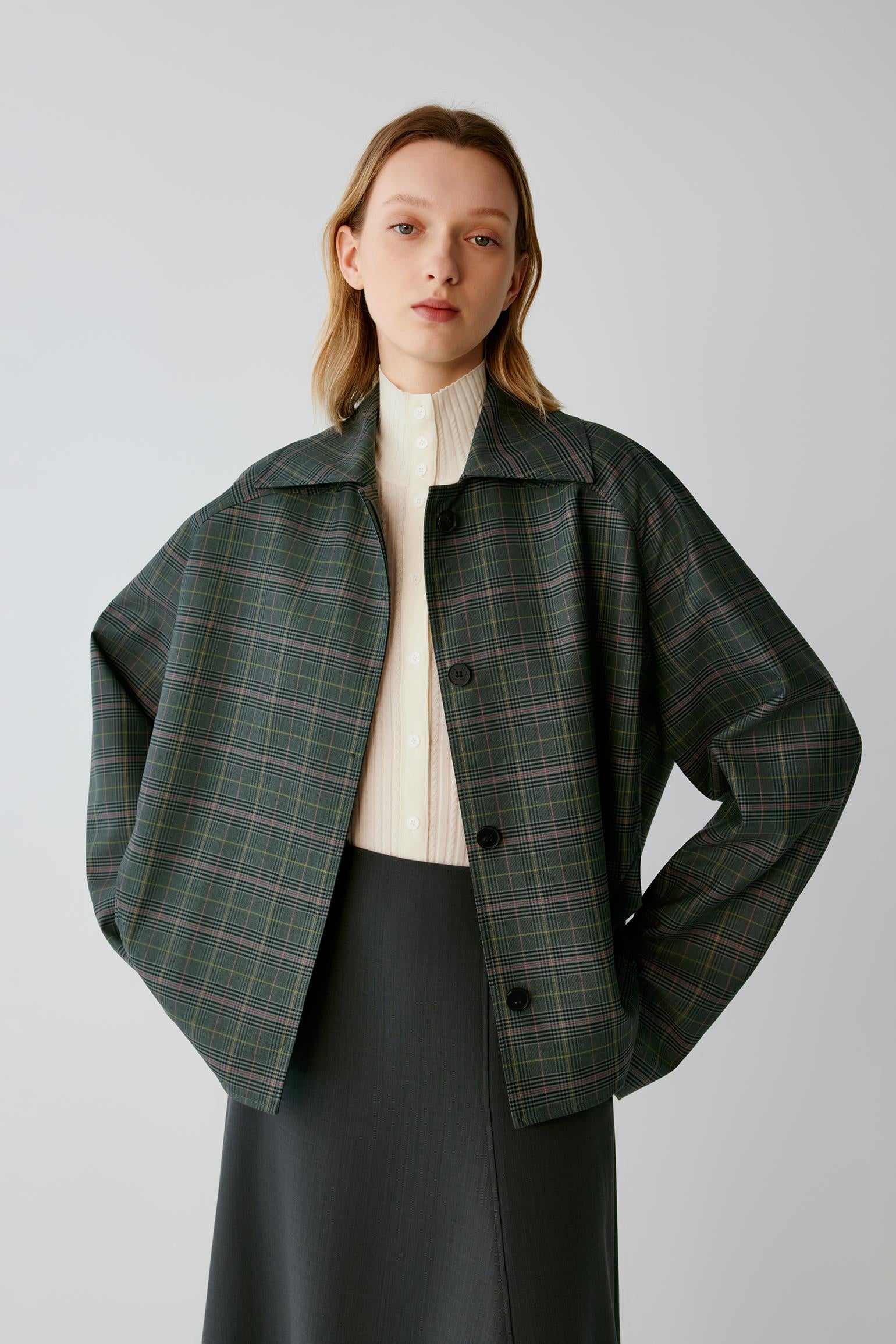 A model wearing a dark green plaid cropped jacket over a cream ribbed turtleneck and a dark skirt, photographed against a neutral light gray studio background.
