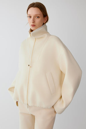 Front view of a woman wearing a cream wool cashmere jacket, showing the relaxed silhouette, high stand collar and two-way front zip fastening.