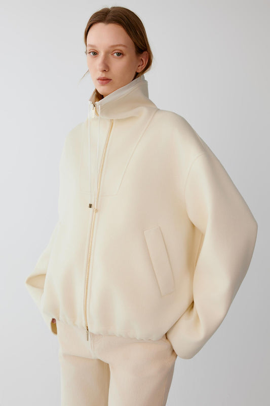 Front view of a woman wearing a cream wool cashmere jacket, showing the relaxed silhouette, high stand collar and two-way front zip fastening.