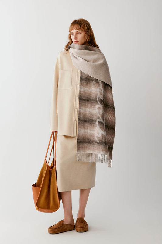 A model is shown from the back wearing a cream-colored coat and skirt, with a large beige and brown striped scarf draped over her shoulders. She is holding a large tan leather tote bag and wearing brown suede loafers, set against a plain light gray studio background.