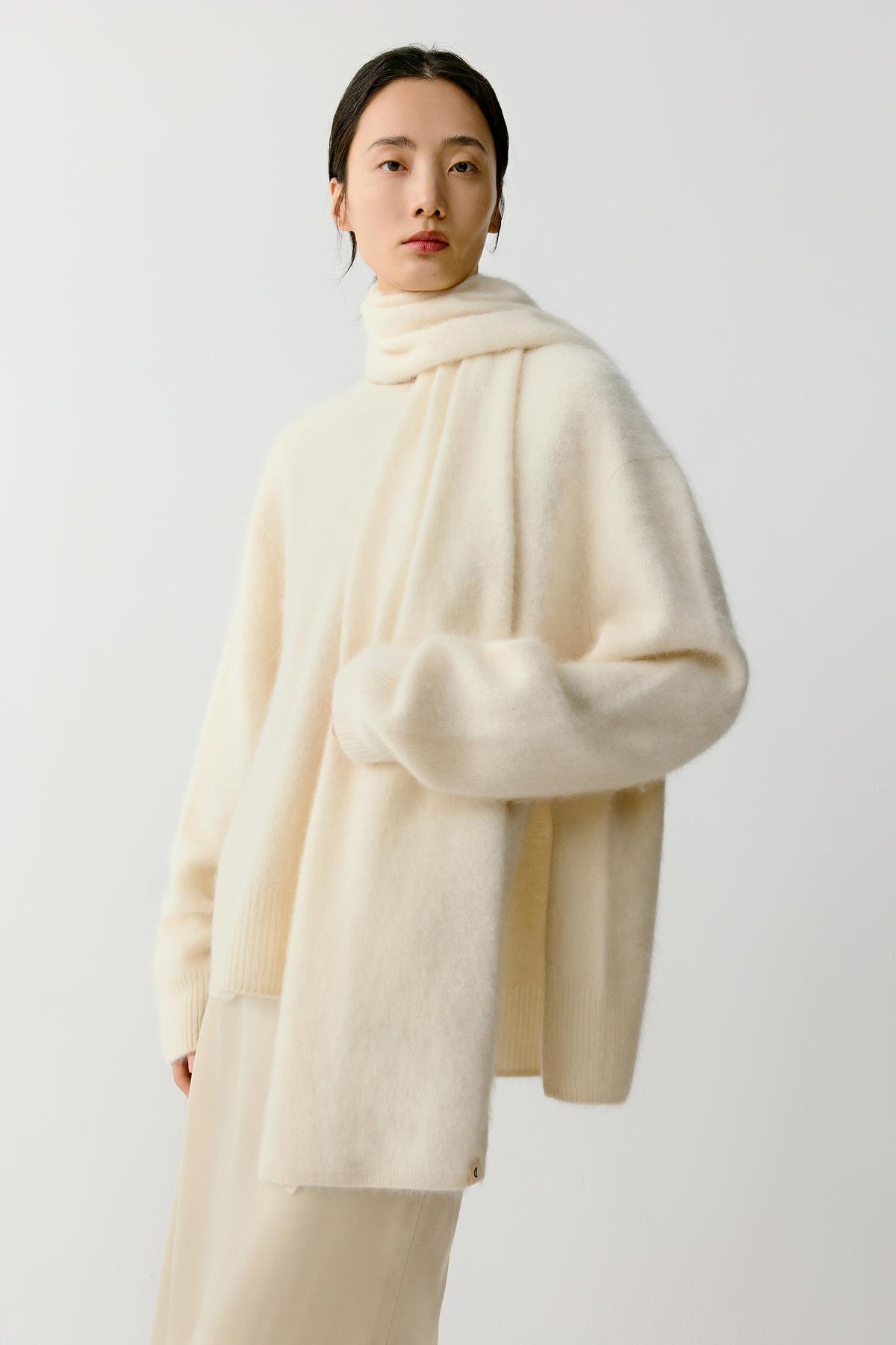 Full-length front view showing the scarf layered over a tonal outfit. The lightweight yet voluminous cashmere falls naturally through the body, demonstrating its soft structure and versatility as a layering piece. The monochrome ivory palette emphasizes the understated character of the accessory.