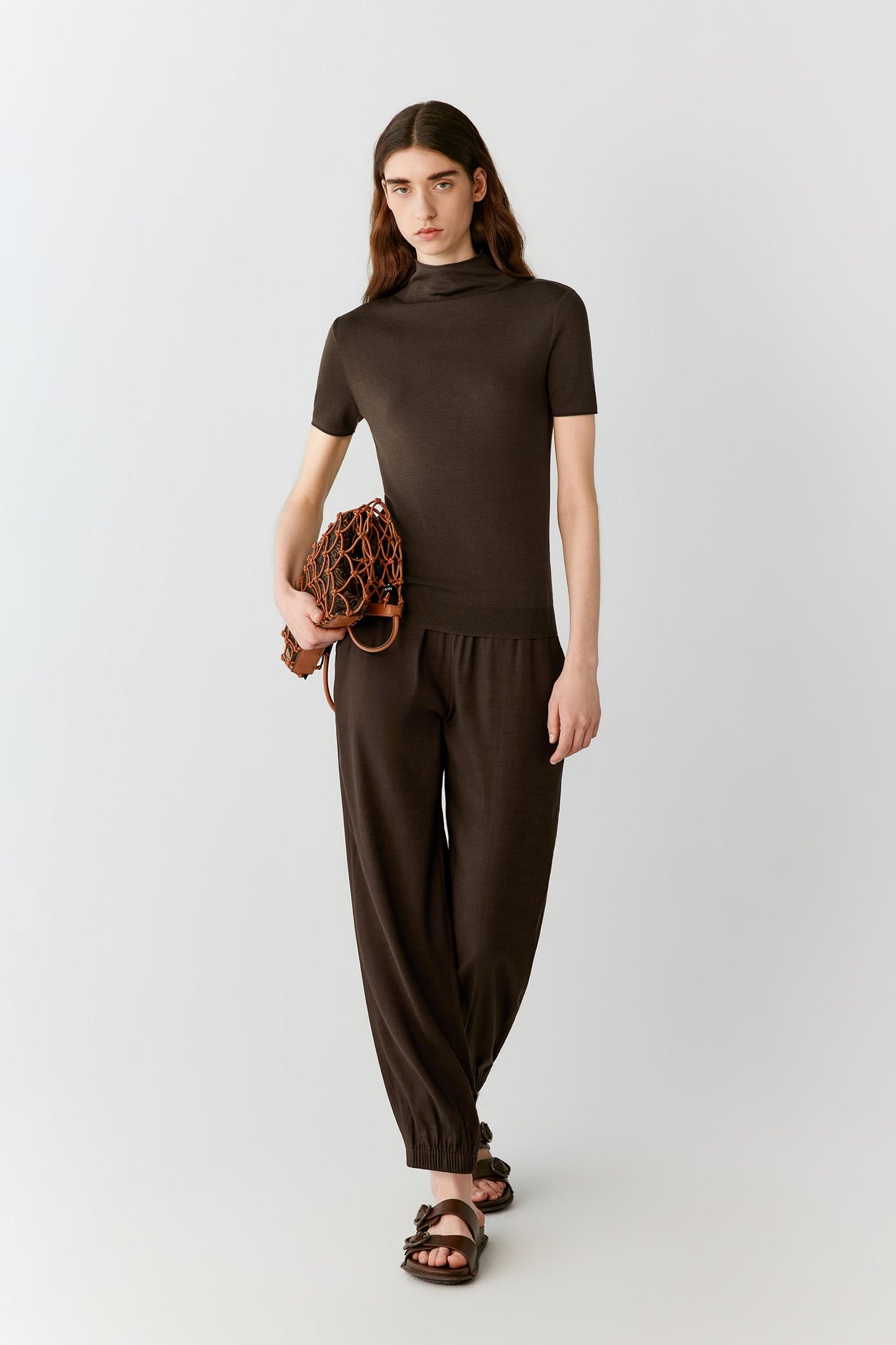 Front view of a woman wearing a dark brown short-sleeved top in fine-gauge silk yarn, highlighting the fitted silhouette, lightweight knit construction and smooth, compact surface. The mock neckline, short sleeves and clean lines create a refined, minimalist appearance.