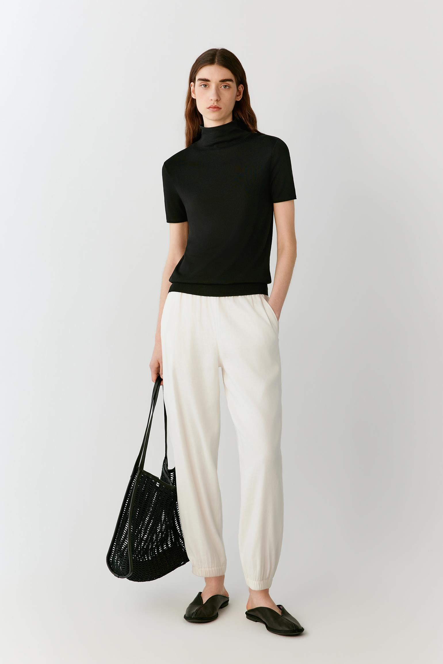Front view of a woman wearing a deep black short-sleeved top in fine-gauge silk yarn, highlighting the fitted silhouette, lightweight knit construction and smooth, compact surface. The mock neckline, short sleeves and clean lines create a refined, minimalist appearance.
