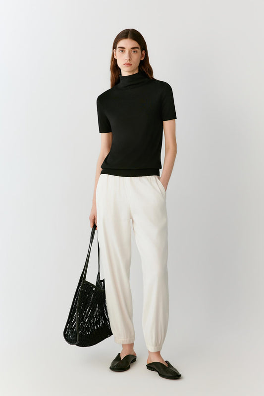 Front view of a woman wearing a deep black short-sleeved top in fine-gauge silk yarn, highlighting the fitted silhouette, lightweight knit construction and smooth, compact surface. The mock neckline, short sleeves and clean lines create a refined, minimalist appearance.