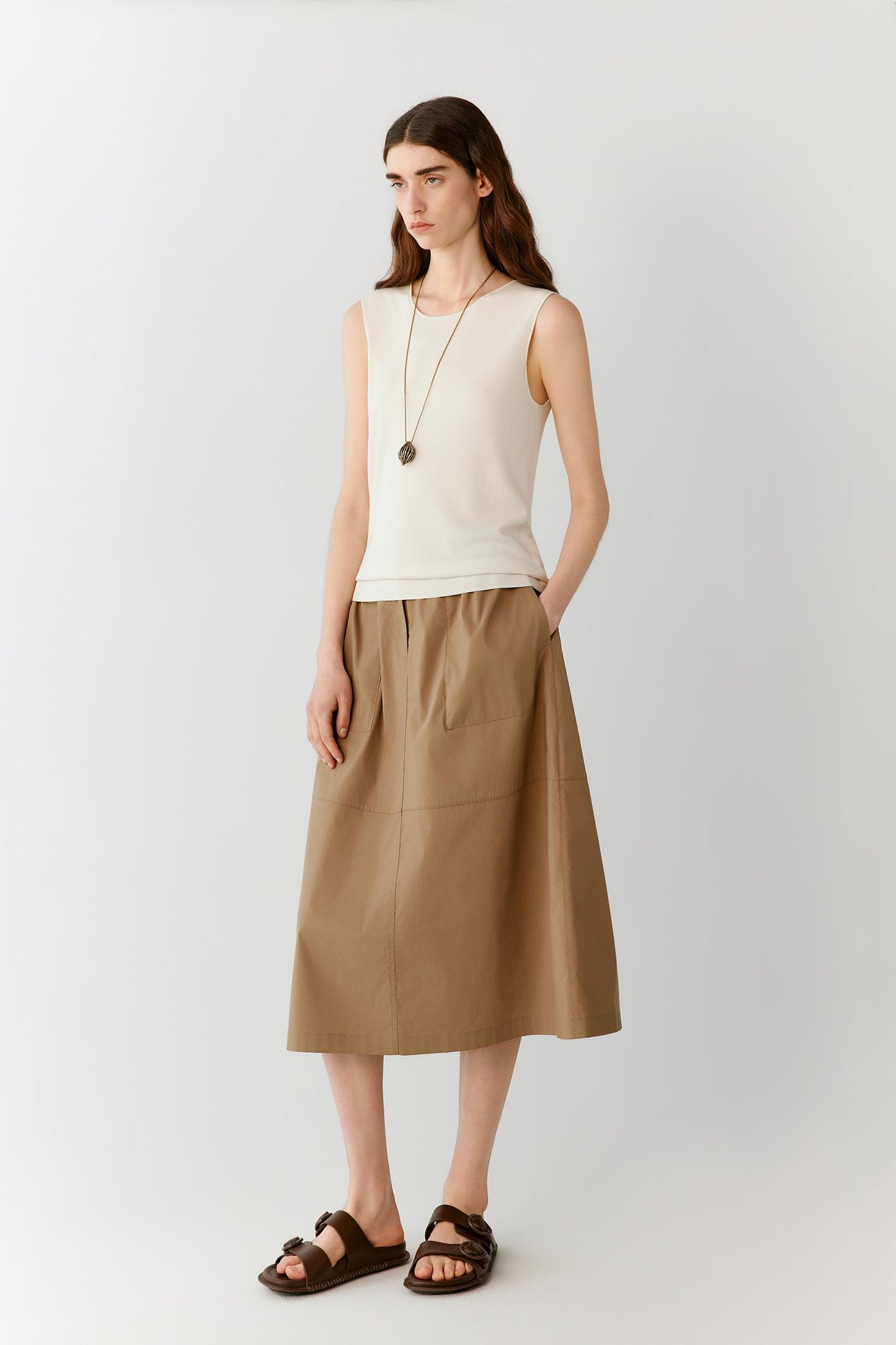 Front view of a woman wearing a brown skirt in a cotton blend, highlighting the fitted waistband, softly flared silhouette and lightweight construction. The smooth surface and subtle tonal variations of the fabric create a clean, understated appearance with fluid movement through the hem.