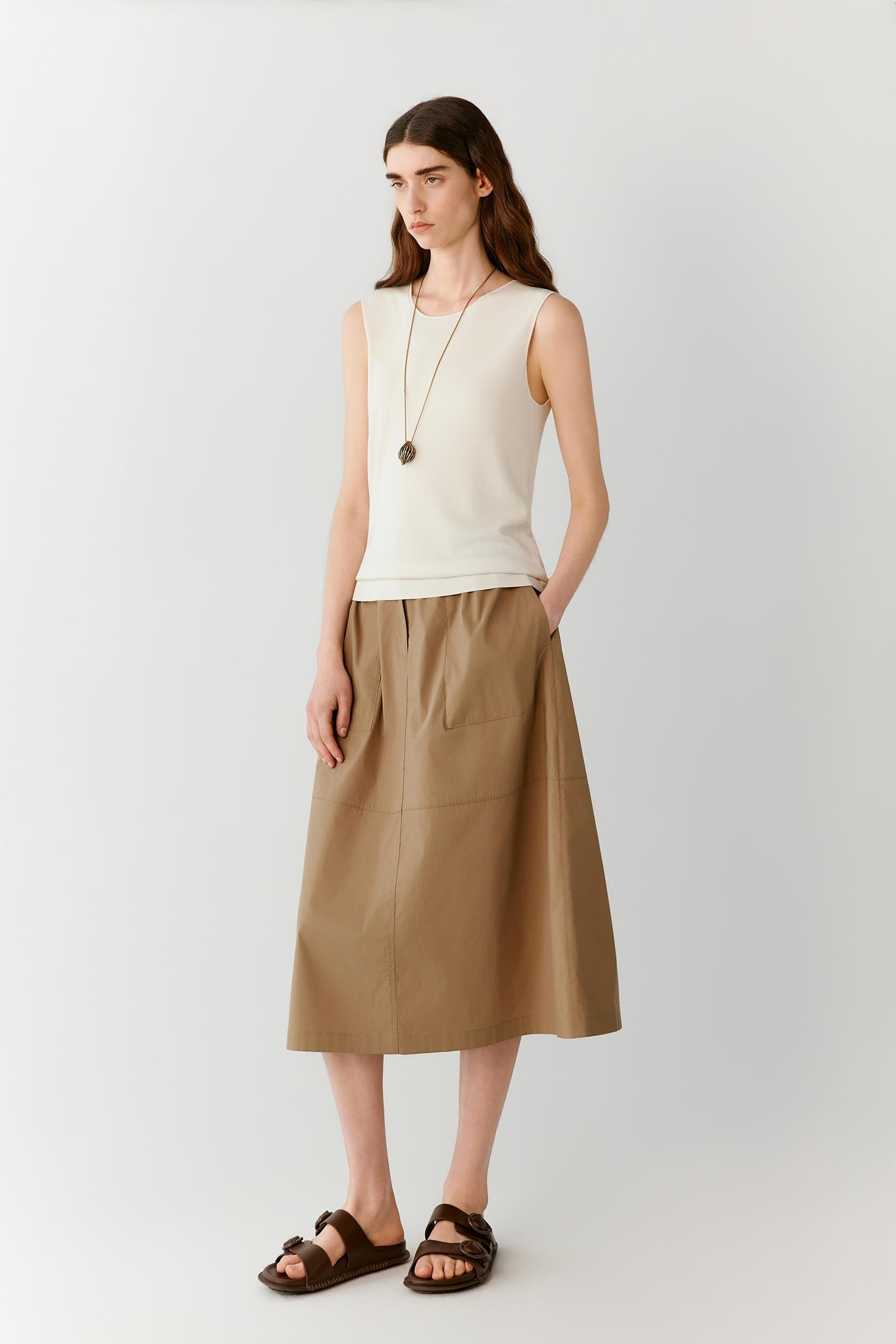 Front view of a woman wearing a brown skirt in a cotton blend, highlighting the fitted waistband, softly flared silhouette and lightweight construction. The smooth surface and subtle tonal variations of the fabric create a clean, understated appearance with fluid movement through the hem.