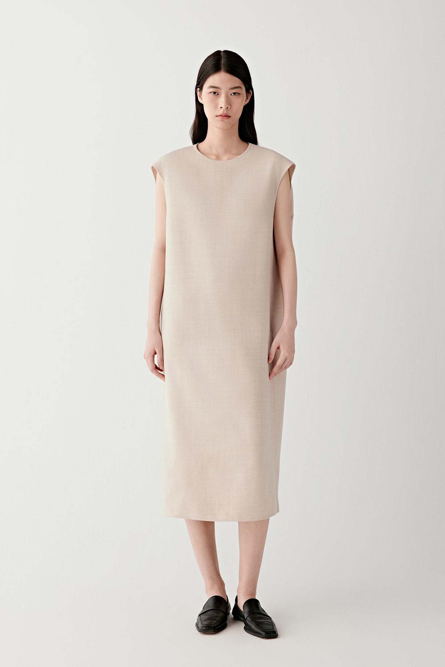 Full-length studio photograph of a woman standing against a light grey background wearing a sleeveless midi dress in lightweight wool in a beige melange shade. The dress has a straight, relaxed silhouette that falls below the knees to mid-calf length. It features a round neckline, clean armholes without sleeves and discreet side pockets integrated into the side seams. The model stands facing forward wearing black flat shoes.