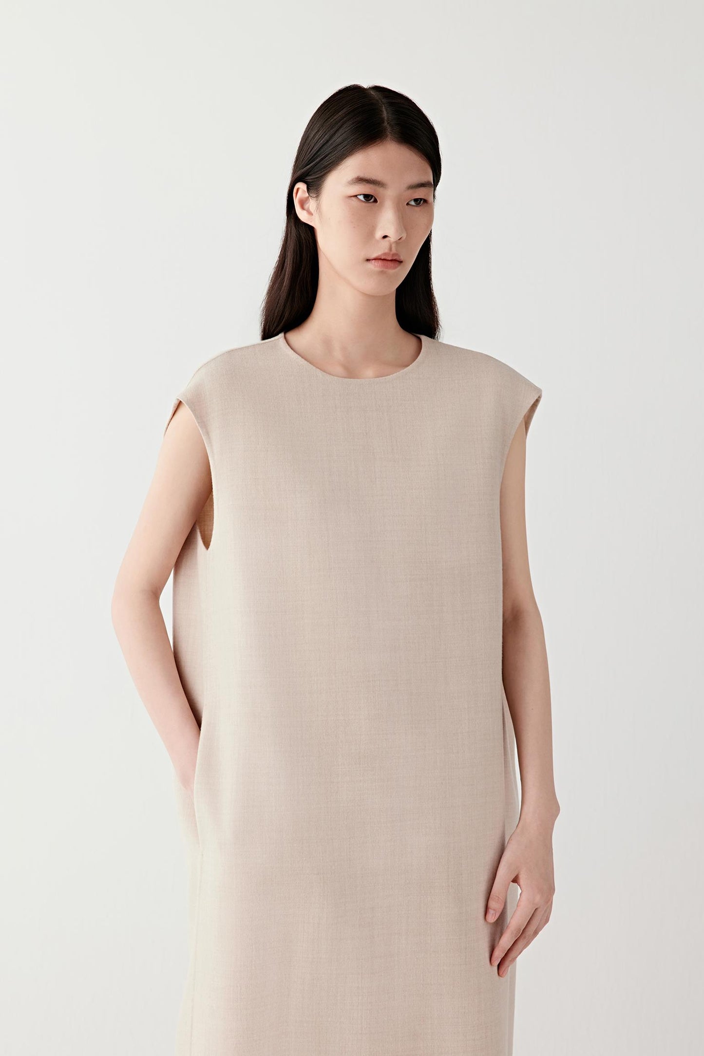 Woman wearing a beige melange lightweight wool dress with a straight, sleeveless cut; mid-length front view highlighting the fluid drape, clean lines and soft structure through the body.