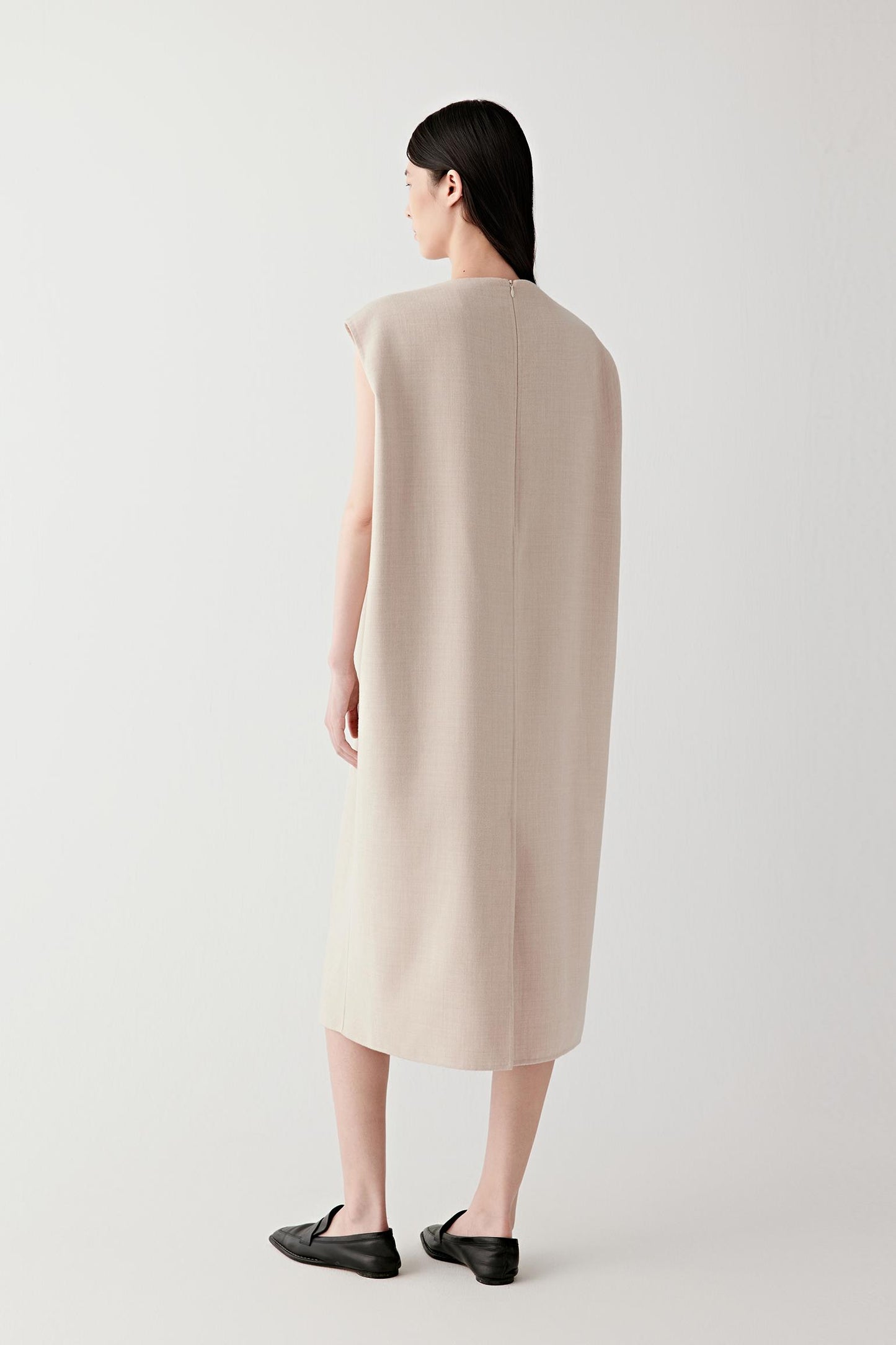 Back view of the sleeveless beige wool dress showing the straight silhouette, smooth back panel construction and the mid-calf length of the garment.