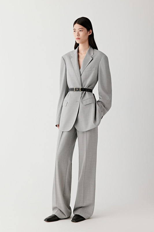 Full-length front view of the model standing, highlighting how the slim leather belt sits at the natural waist over a structured blazer, emphasizing proportion, clean lines and contrast between the black leather and light fabric