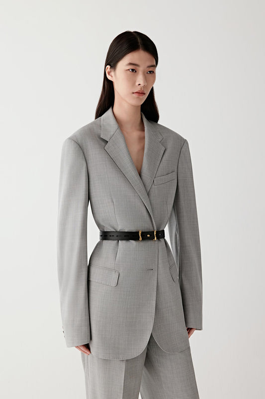 Three-quarter front view of a woman wearing a light grey tailored suit cinched at the waist with a slim black calf leather belt, showing the narrow width, smooth surface and how the belt defines the silhouette, with the antique gold-tone brass buckle positioned centrally