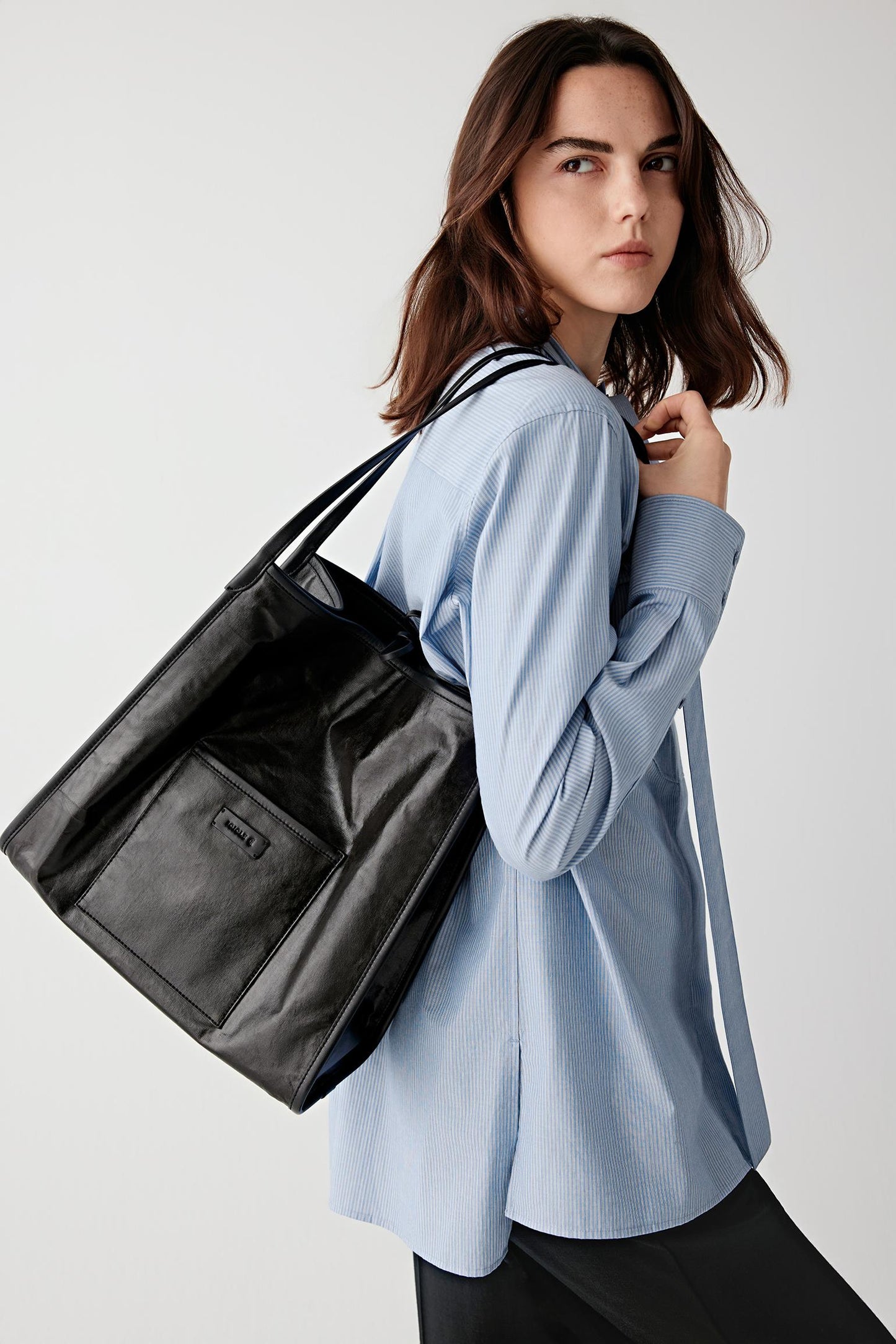 Three-quarter back view of a woman carrying a large black leather tote bag over a light blue shirt, showing smooth surface, structured volume and shoulder positioning