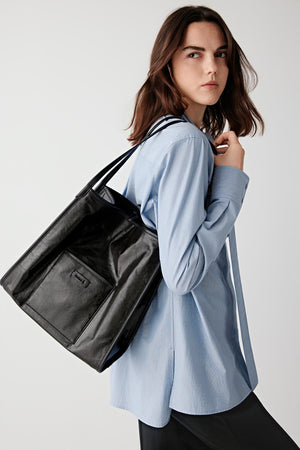 Three-quarter back view of a woman carrying a large black leather tote bag over a light blue shirt, showing smooth surface, structured volume and shoulder positioning
