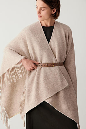 Three-quarter front view of a woman wearing a layered outfit with a soft beige wrap-style top over a dark inner layer, cinched at the waist with a slim brown calf leather belt, showing the narrow width, smooth leather surface and how the belt structures the silhouette while creating a natural contrast with the flowing fabric
