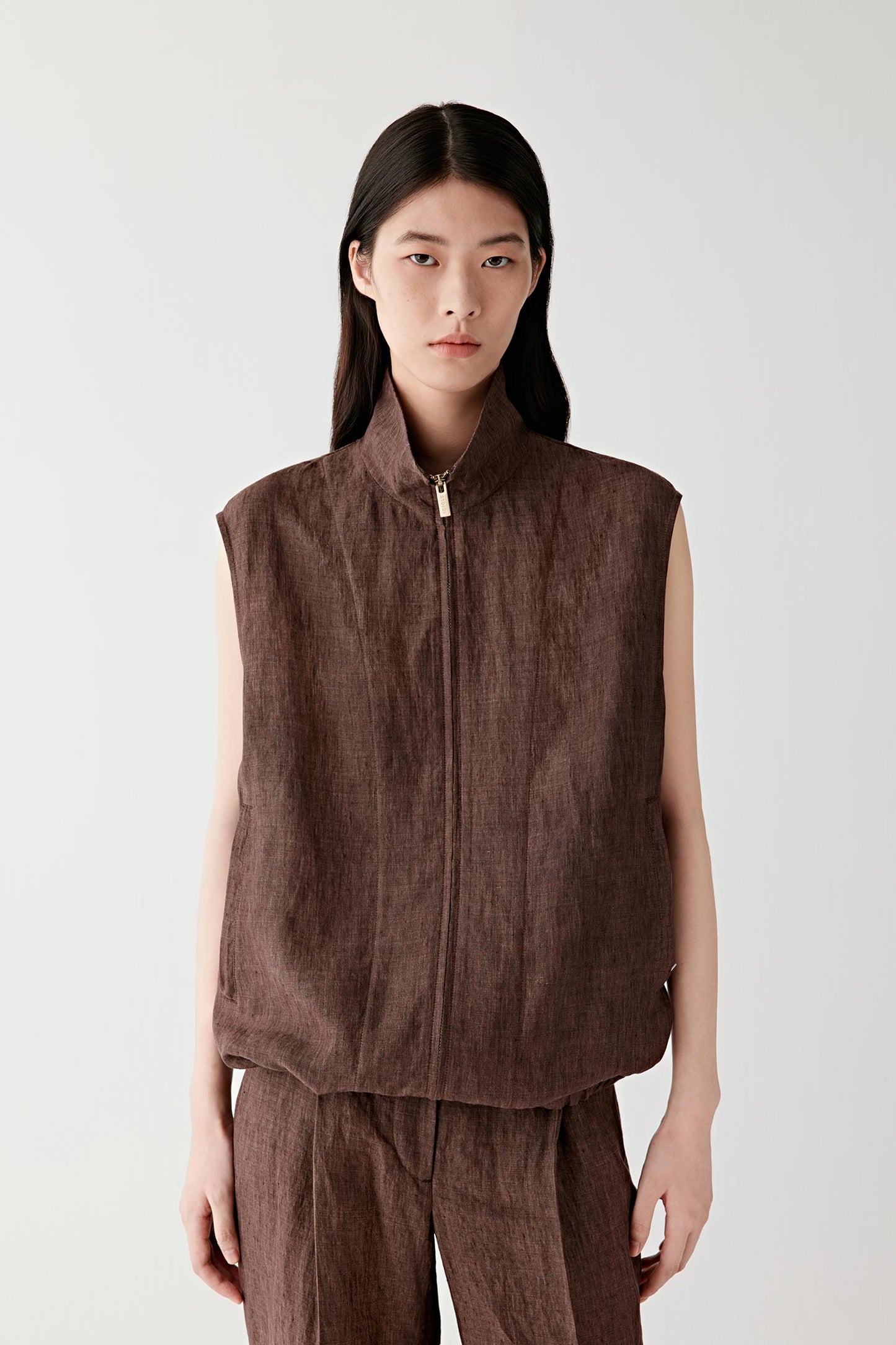 Three-quarter front view of a woman standing slightly angled wearing a brown sleeveless linen vest with a front zip closure, structured shoulders and a fitted silhouette, showing the textured surface and clean lines along the torso