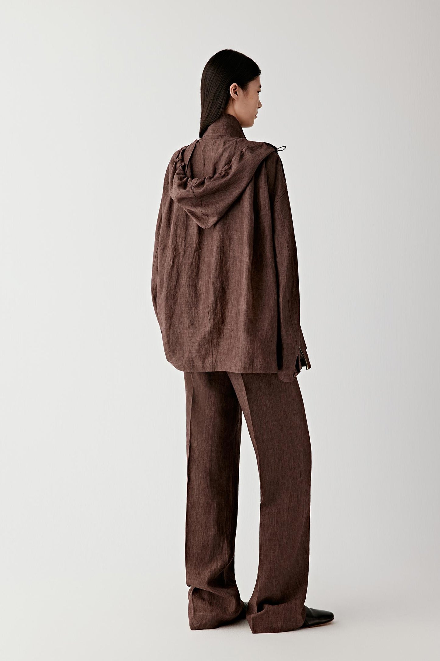 Full-length back view showing the hood laid flat against the upper back, the straight cut through the body and the natural fall of the linen fabric from shoulders to hem
