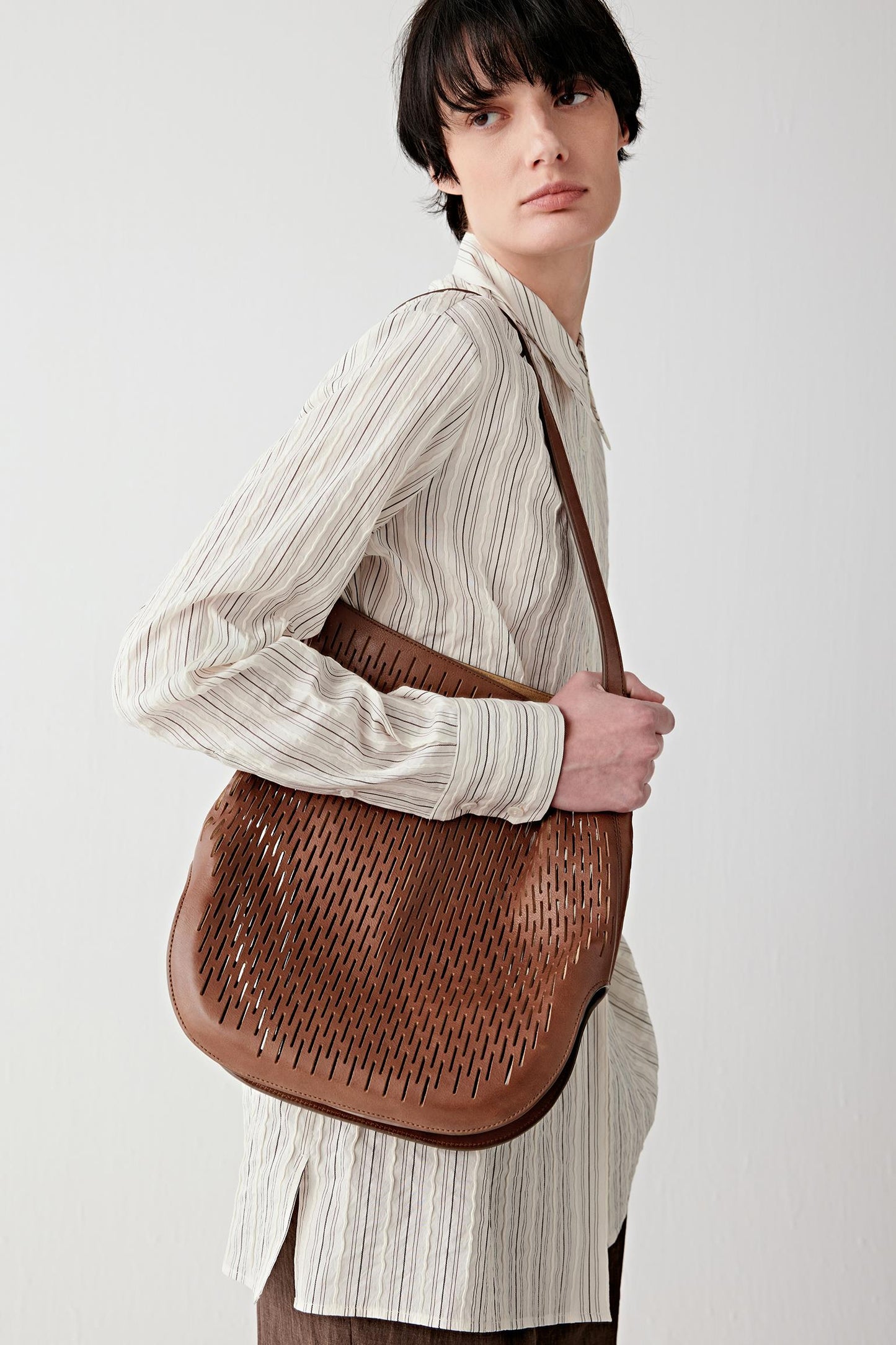 Woman wearing a dark brown perforated leather hobo bag with a soft slouchy silhouette, carried on the shoulder, full-length view highlighting the curved shape and supple drape of the leather.