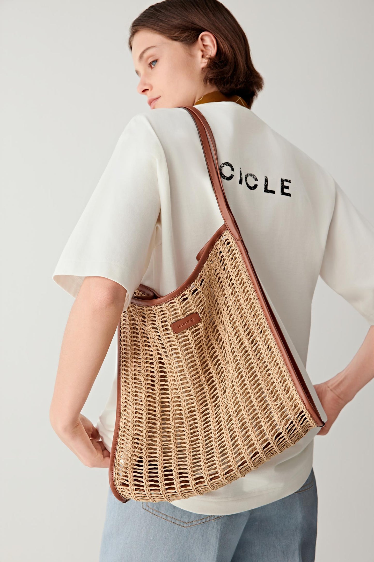 Back three-quarter view of a woman carrying a beige cotton mesh tote bag with leather shoulder straps, worn over a white top, highlighting the open mesh construction, transparency and soft flexible structure